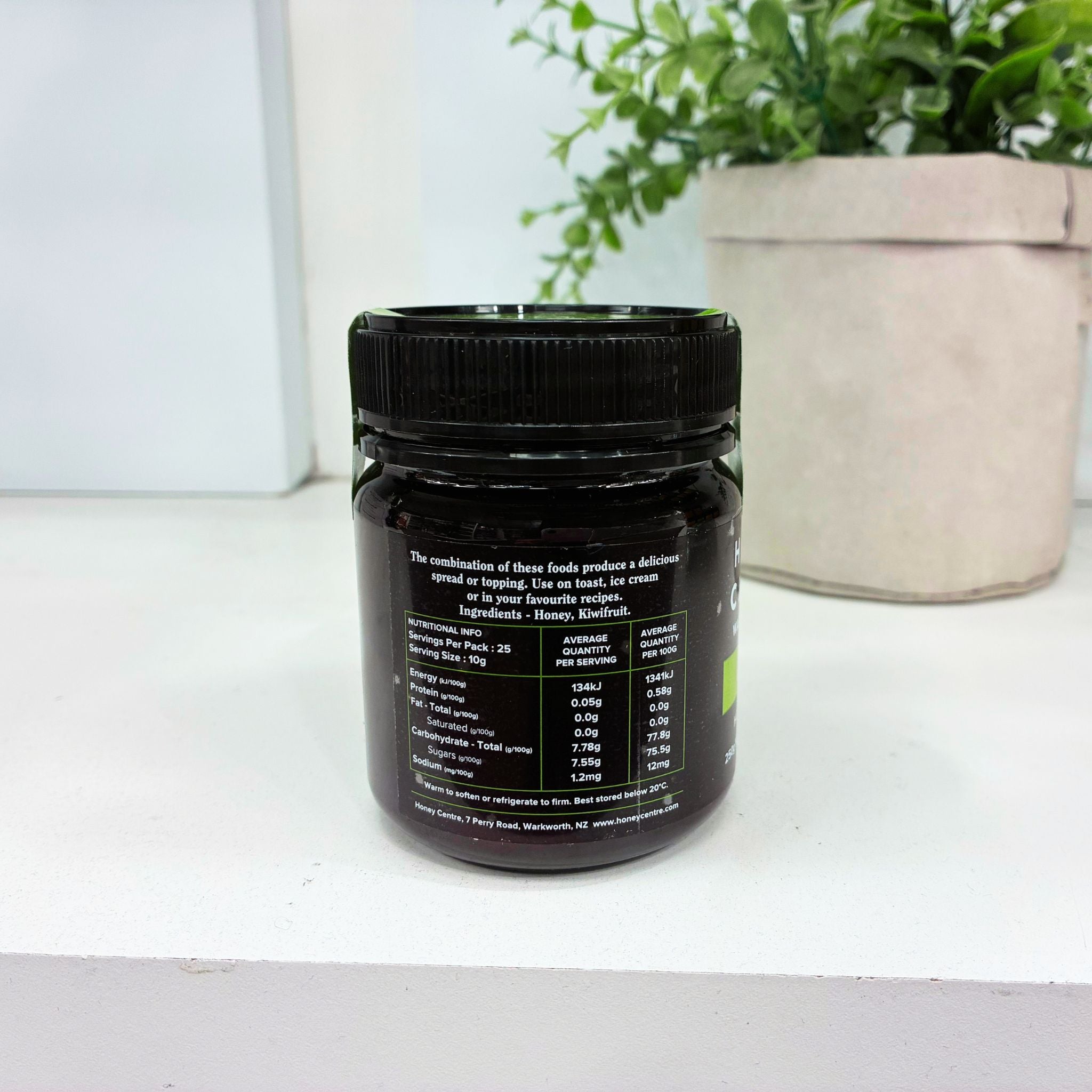 A dark jar of kiwifrit honey with a black lid, showing the back label with nutritional information, set on a white surface with a potted plant in the background.