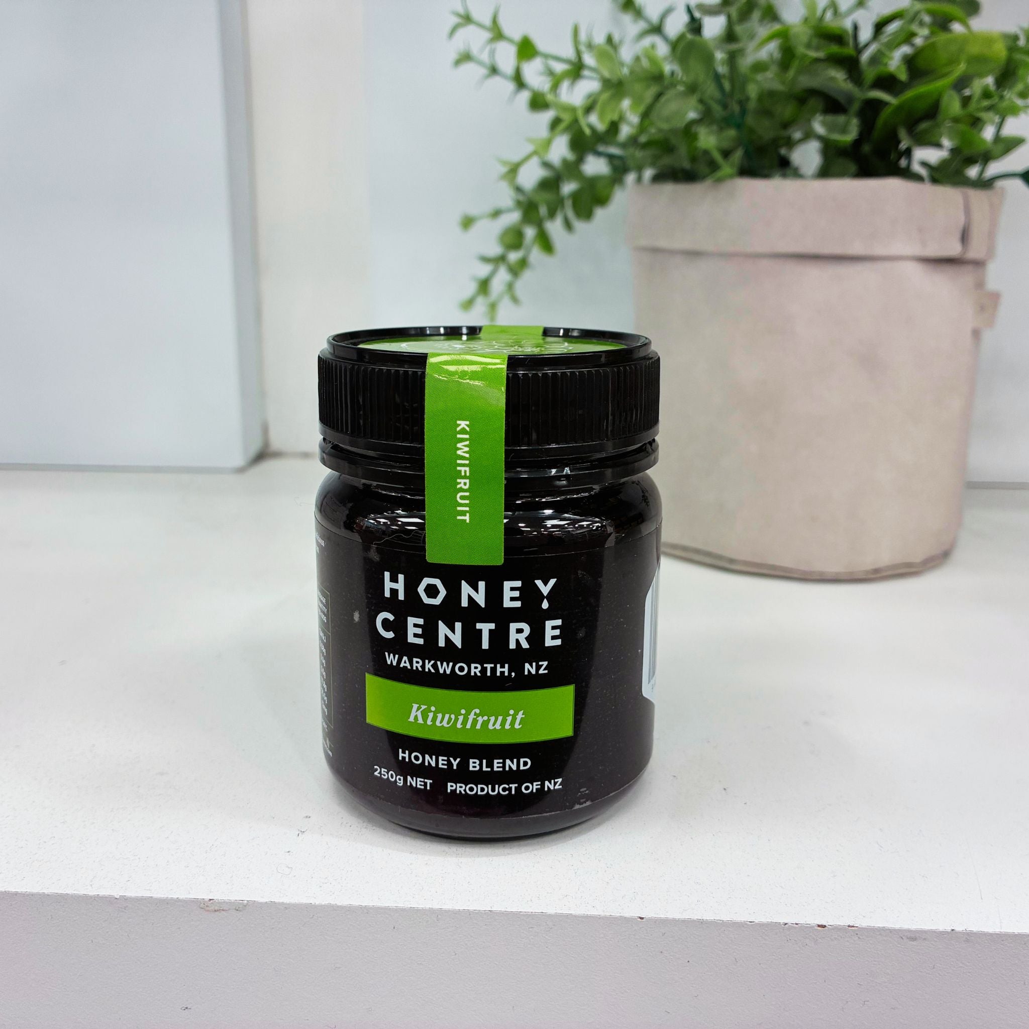 A jar of Honey Centre kiwifruit honey blend on a white shelf with a potted plant in the background. The jar is dark with a black lid and features a bright green label and a vertical green seal.