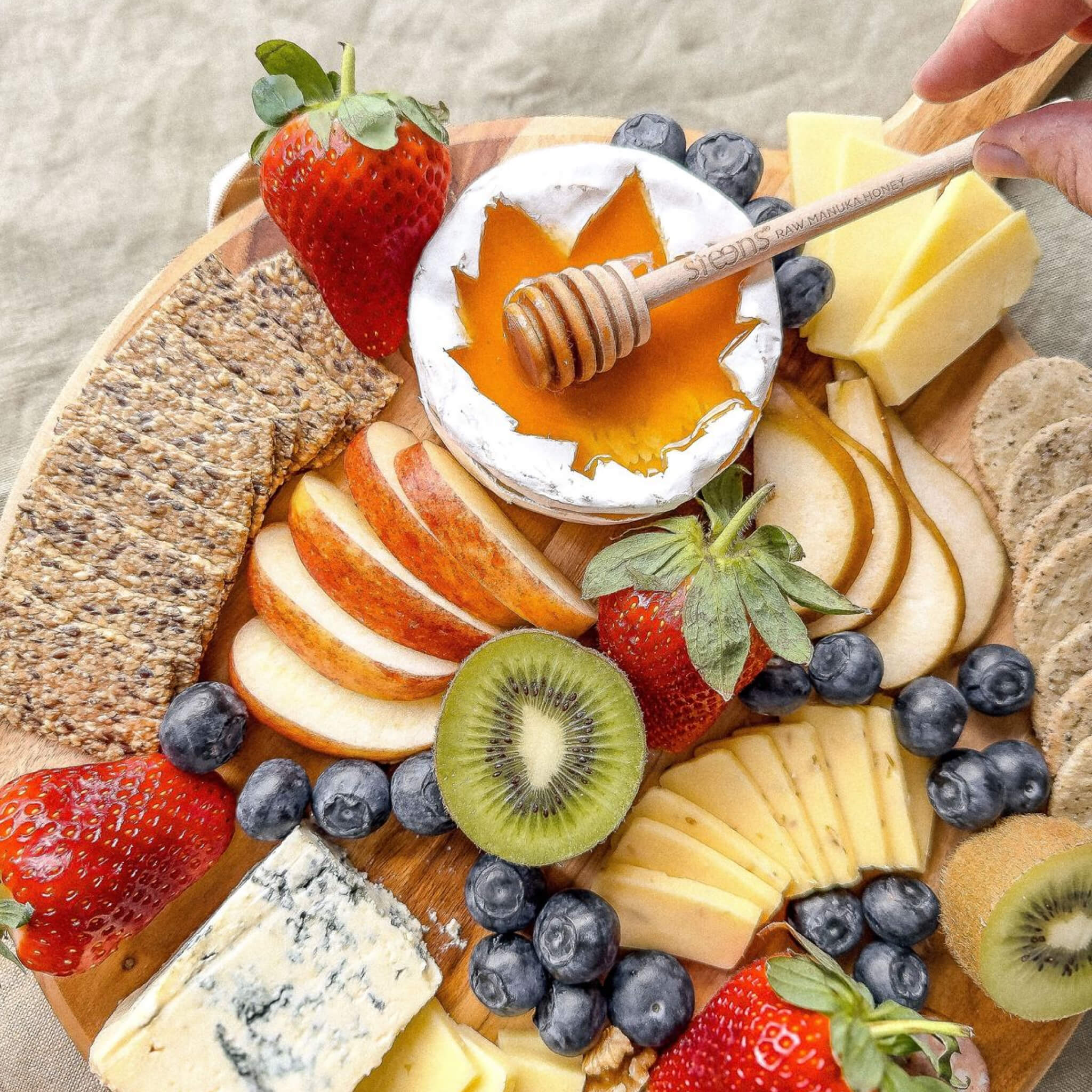 Assorted cheeses, fruits, and crackers on a wooden board with Steens Manuka honey.