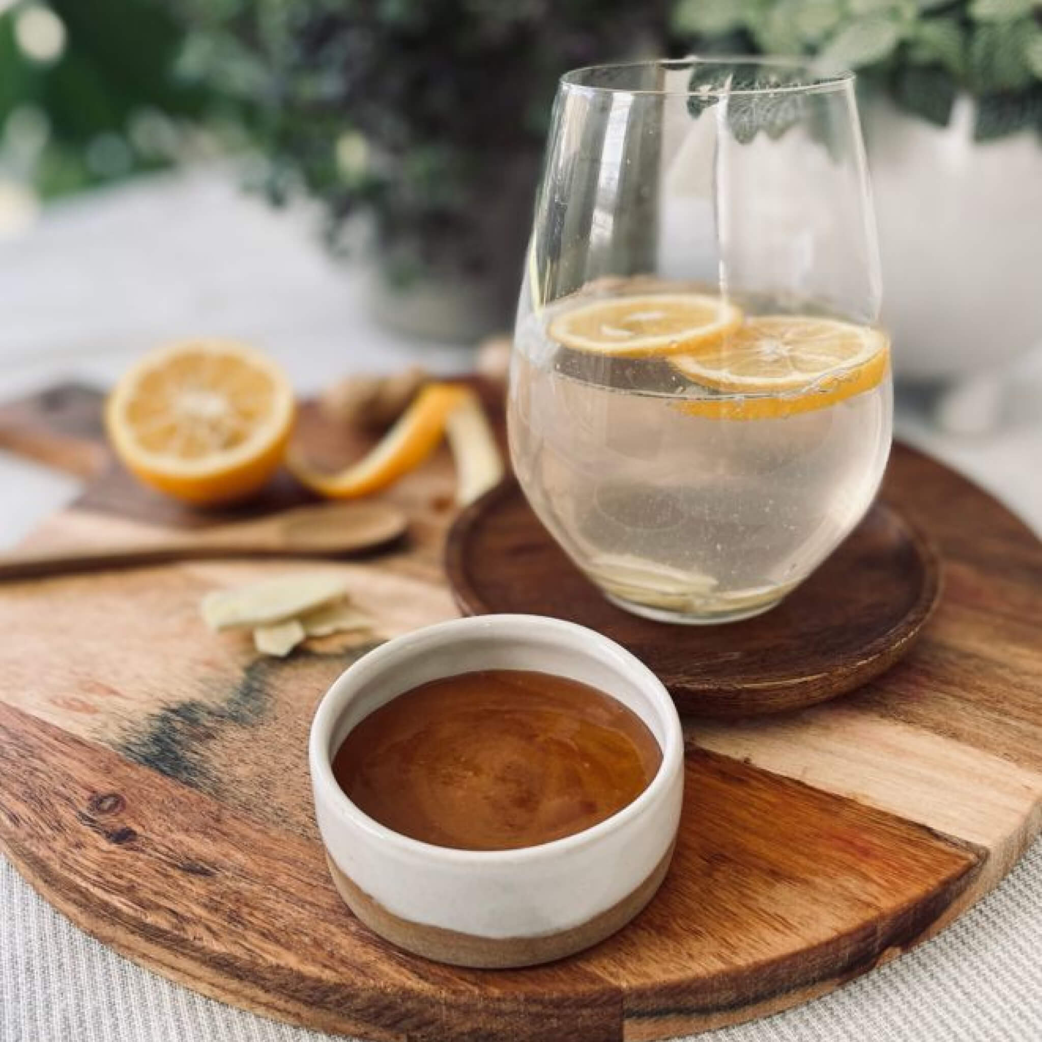 Glass of water with lemon slices on a wooden coaster, accompanied by a small bowl of Steens Manuka Honey on a wooden cutting board.