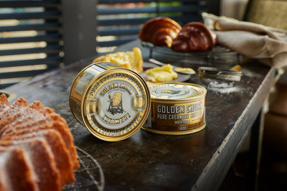 Two tins of Golden Churn Pure Creamy Butter on a rustic wooden surface with baked goods.