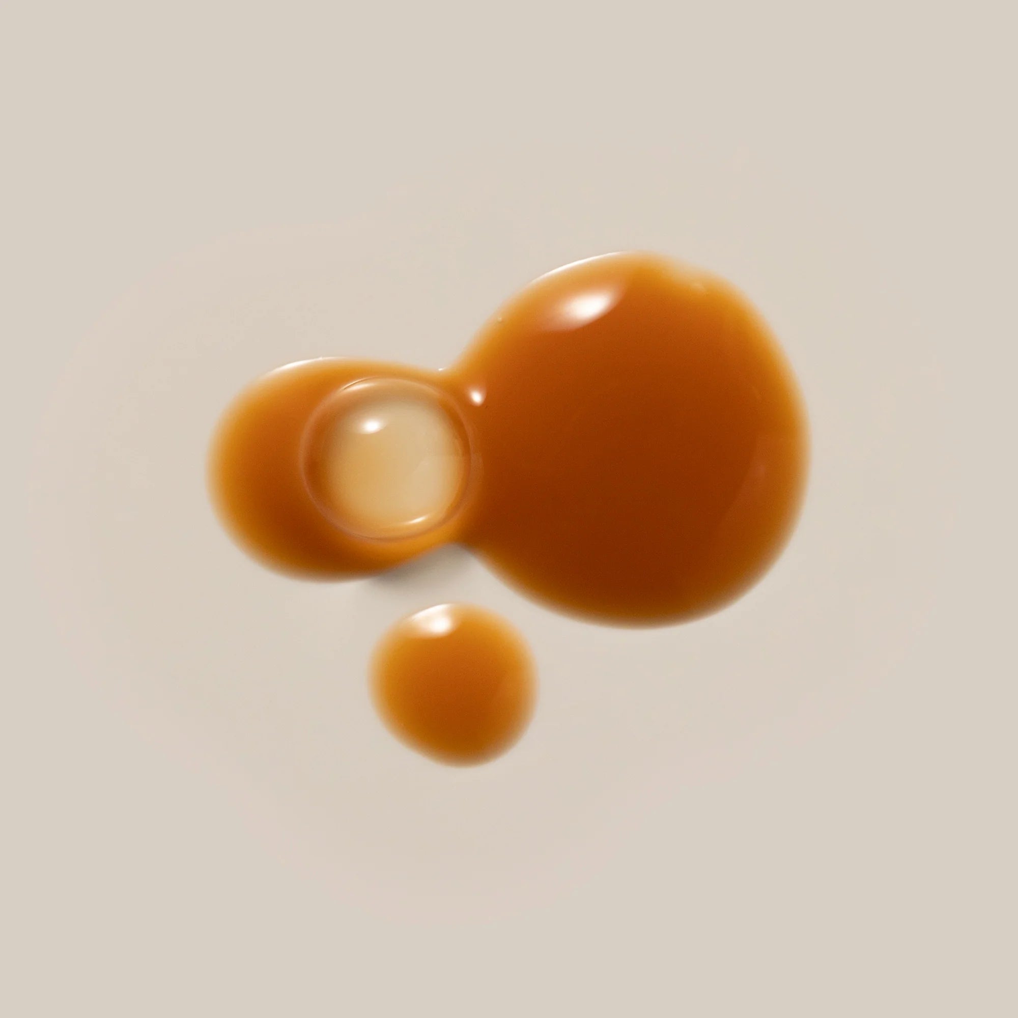 Three droplets of caramel-colored liquid of Antipodes Hosanna on a beige background