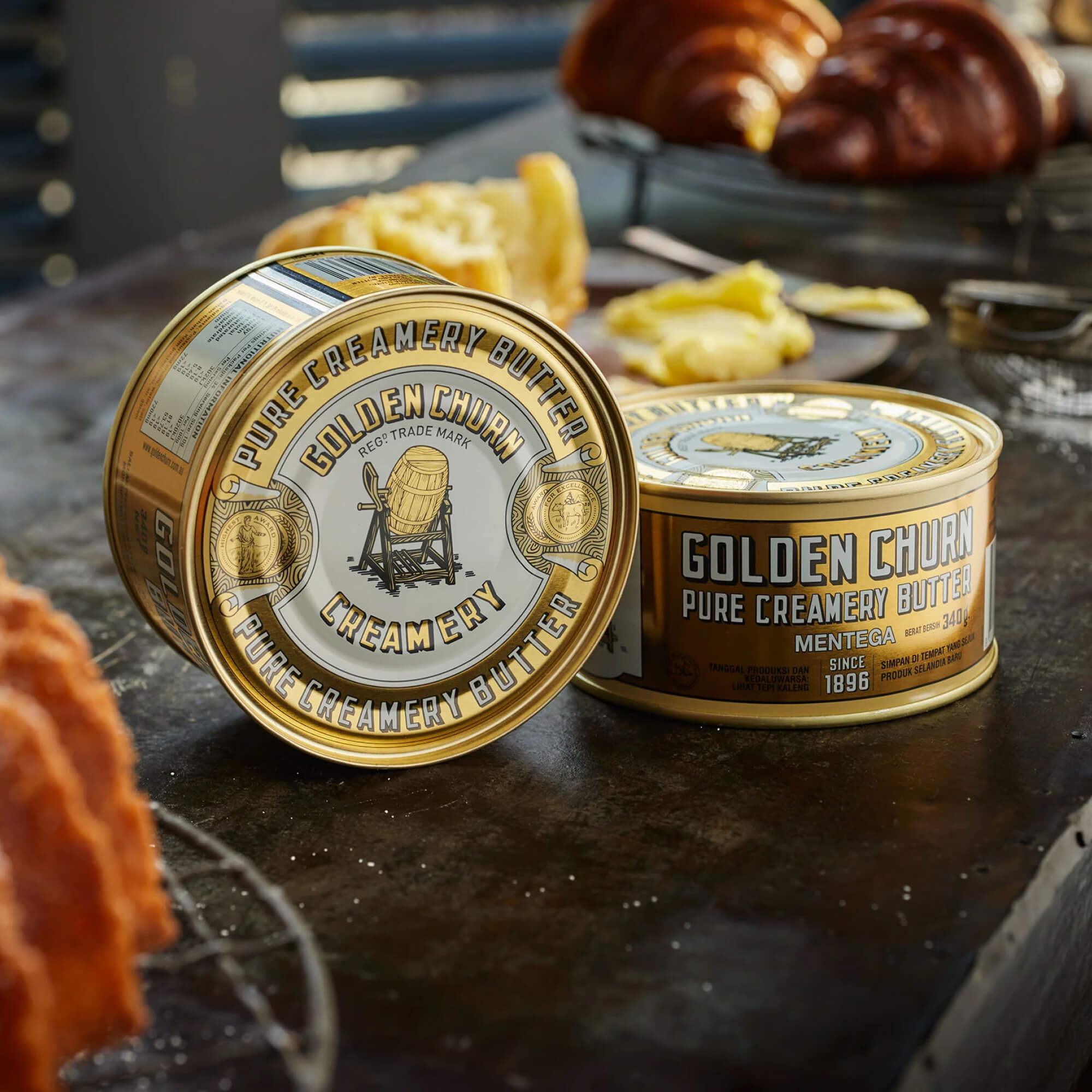Two tins of Golden Churn butter on a rustic surface with food items in the background.