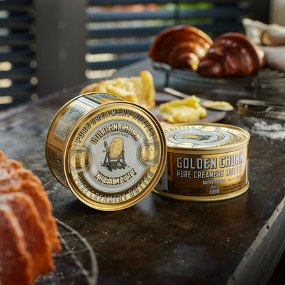Two tins of Golden Churn Pure Creamy Butter on a rustic wooden surface with baked goods.