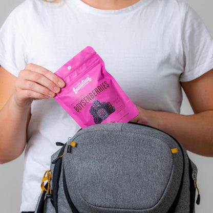 Person holding a pink package labeled Little Beauties 'Boysenberries' next to a gray bag on a plain background