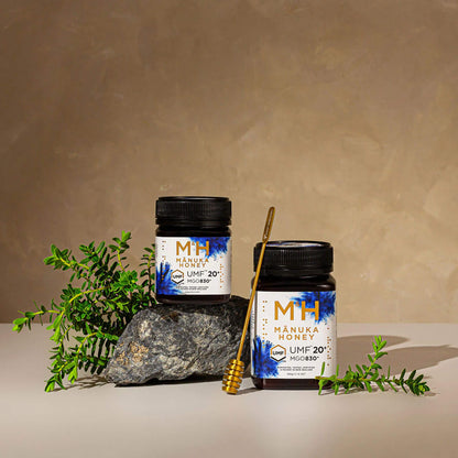 Two jars of M&H Manuka Honey UMF20+ 250g and 500g with a background of greenery and a neutral wall.