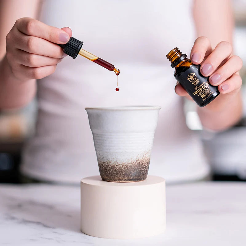 Person pouring Manuka South propolis extract from a dropper bottle into a small cup on a white surface.