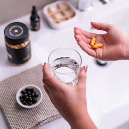 Person holding a glass of water with two yellow pills, surrounded by health supplements on a white surface.