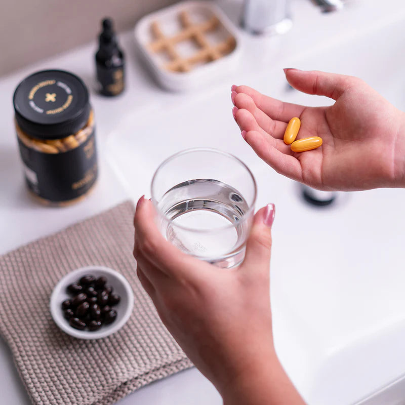 Person holding a glass of water with two yellow pills, surrounded by health supplements on a white surface.