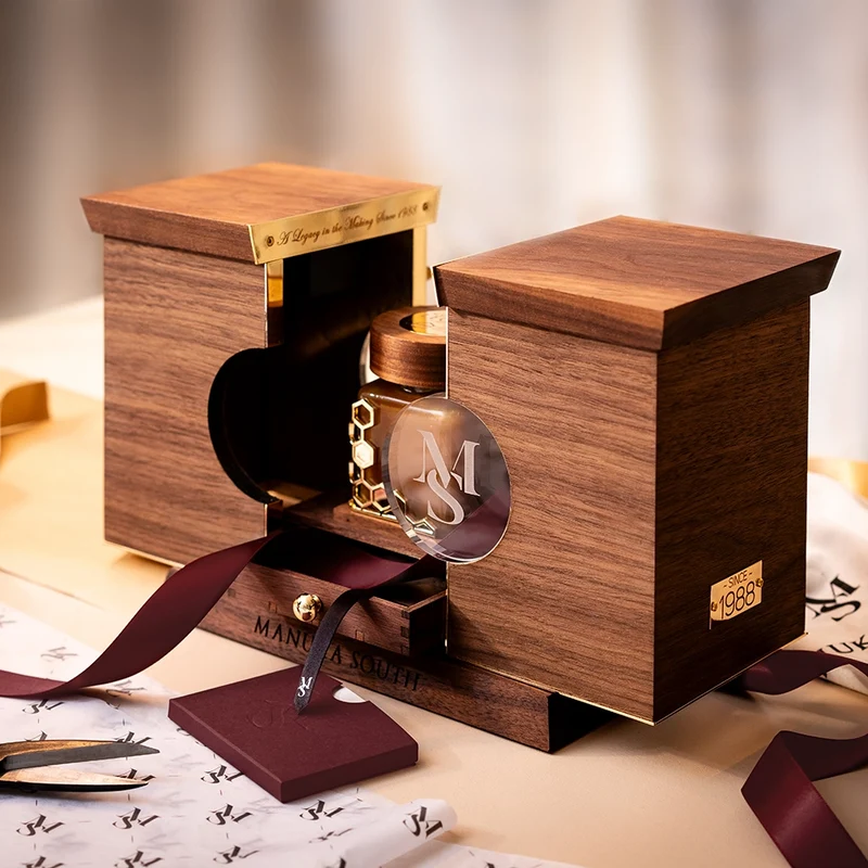 A luxury Manuka Honey jar with a clear glass insert and gold details, presented in a custom wooden display box with a dark burgundy ribbon, on a beige surface with branded papers.