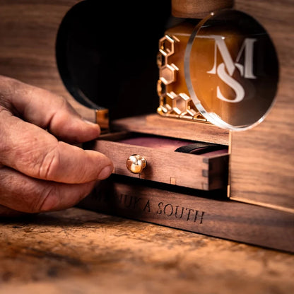 A close-up of a hand opening a wooden drawer of a personalized Manuka South Manuka Honey box. The box is made of dark wood with gold metal accents, including a pull knob and a honeycomb pattern on the bottle cap.