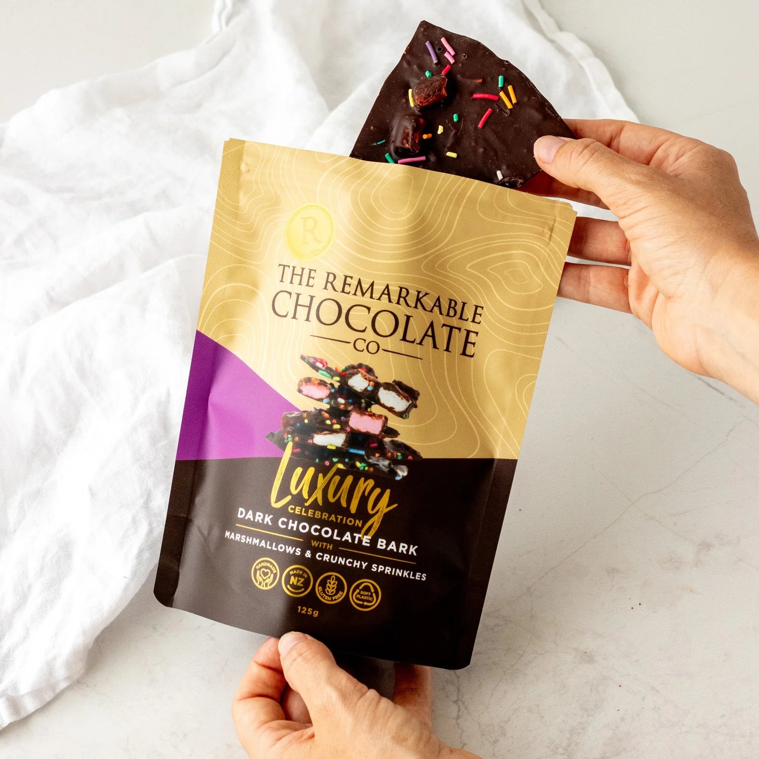 Hand holding a package of The Remarkable Chocolate Co. Luxury Dark Chocolate Bark mashmallow and sprinkles with a piece of chocolate bark on top.
