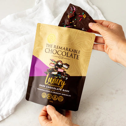 Hand holding a package of The Remarkable Chocolate Co. Luxury Dark Chocolate Bark mashmallow and sprinkles with a piece of chocolate bark on top.